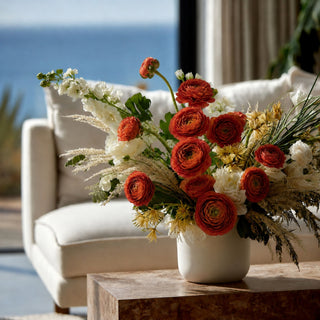 A vibrant floral arrangement featuring red ranunculus, white stock, and yellow wildflowers in a white ceramic vase, styled on a wooden table in a bright living room with a white armchair and a view of the ocean in the background.