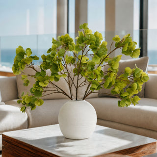 A green leafy branch arrangement in a round white textured ceramic vase, placed on a marble-topped coffee table in front of a beige sofa with large windows.