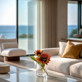 A modern living room with a clear glass vase holding two orange-red flowers on a marble coffee table, with a white sofa, yellow pillow, and large windows overlooking the ocean in the background.