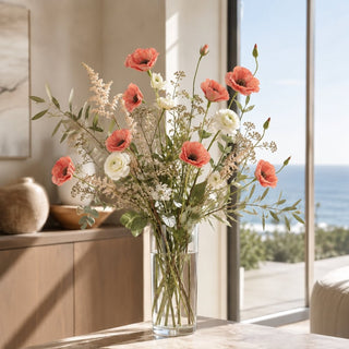 A floral arrangement in a clear glass vase on a table, featuring orange bellflowers, white ranunculus, and green foliage, set in a bright room with a window overlooking the ocean.