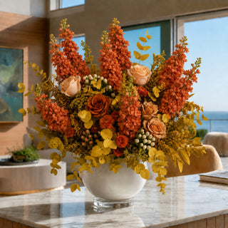 A large, vibrant autumn floral arrangement in a white ceramic bowl vase, set on a marble countertop in a modern, sunlit interior with a view of the ocean.
