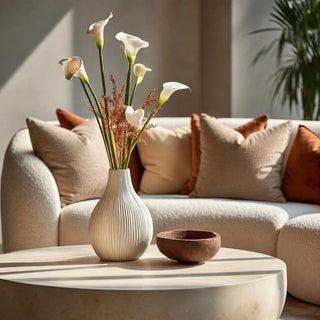 A ribbed ceramic vase filled with white real touch fake calla lilies and dried stems sits on a round stone coffee table in a modern living room with a beige sofa and cushions.