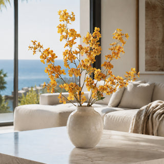 Yellow artificial flowers arranged in a textured cream ceramic vase, placed on a marble coffee table in a modern living room with a large window overlooking the ocean.