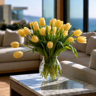 A vibrant bouquet of yellow tulips in a clear glass vase, placed on a marble-topped coffee table in a modern living room setting with a sofa and large windows in the background.