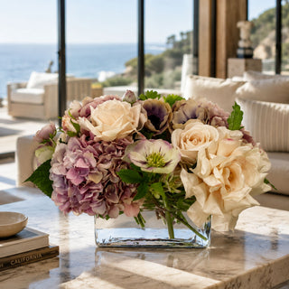 The Montecito Bloom premade True Bloom Real Touch floral arrangement styled in a clear glass vase on a coffee table with cream roses, mauve hydrangeas, soft anemone-style blooms, and green foliage in a refined coastal living room.