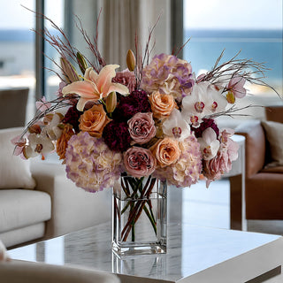 Real touch purple and cream hydrangea stems styled in a luxury faux floral arrangement with peach roses, white orchids, mauve roses, burgundy accent flowers, airy branches, and a clear glass vase in a bright coastal living room.