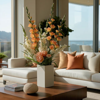Sun-kissed orange True Bloom gladiolus stems styled in a white vase with white florals and peach roses on a wood coffee table in a light-filled living room