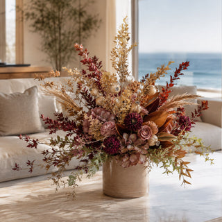Taupe Brown Poppy Pod Spray styled in a fall floral arrangement with burgundy dahlias, dried-style hydrangeas, rust foliage, pampas grass, magnolia leaves, and layered autumn greenery in a ceramic vase on a marble coffee table in a bright coastal living room.