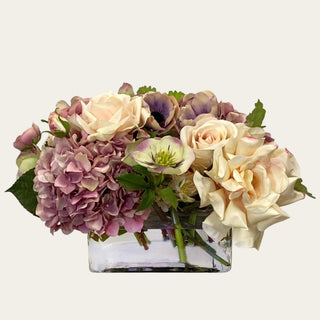 The Montecito Bloom premade True Bloom Real Touch floral arrangement shown in a clear glass vase with cream roses, mauve hydrangea-style blooms, soft lavender accents, and green foliage for year-round tabletop styling.