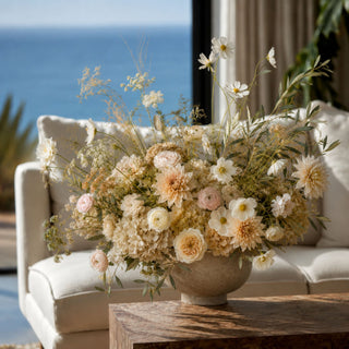 A large, elegant floral arrangement in a textured ceramic pot, styled on a wooden table in front of a cream-colored sofa with a view of the ocean in the background.