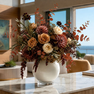 A large, lush dried floral arrangement in a round white ceramic vase, set on a marble countertop in a modern, sunlit interior with a view of the ocean in the background.