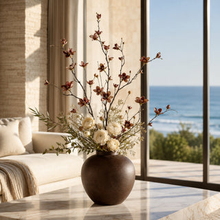 Luxury faux burgundy brown magnolia blossom floral arrangement in a chocolate brown ceramic vase with cream ranunculus, styled on a marble coffee table inside a California coastal home overlooking the Pacific Ocean.