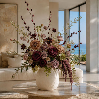 Luxury late summer to early fall faux floral arrangement featuring burgundy velvet willow stems, wine garden roses, taupe dahlias, antique mauve roses, chocolate cosmos, olive branches, and trailing plum amaranthus in a textured white ceramic vase. styled in a bright modern living room.