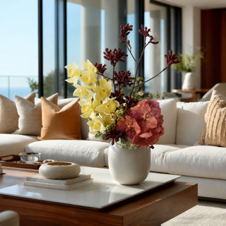 A white ceramic vase filled with a floral arrangement of yellow orchids, pink flowers, and dark burgundy stems, placed on a wooden coffee table in a bright, modern living room with a white sofa and large windows.