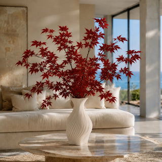 Crimson red faux maple leaf sprays styled in a ceramic vase beside large windows, creating a full fall foliage arrangement in a modern interior.