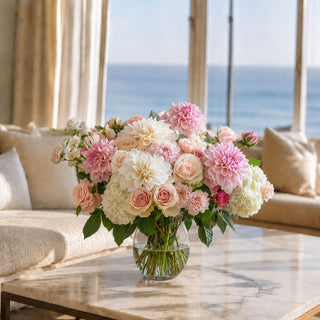 Light pink real touch dahlia stems styled in a glass vase with cream roses, white hydrangeas, blush florals, greenery, and soft garden accents in a bright living room arrangement for spring, summer, and fall styling.