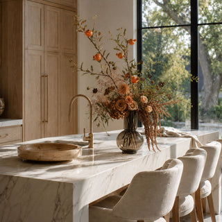 A modern kitchen island with a white marble countertop, a matte brass faucet, a wooden bowl, and a large vase of dried orange flowers, set in a bright kitchen with light wood cabinets and upholstered bar stools.