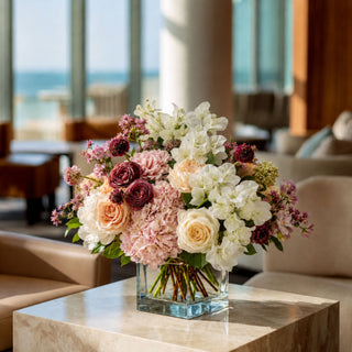 Premium artificial floral arrangement by True Bloom Floral with realistic white bougainvillea, blush hydrangeas, ivory roses, mauve garden roses, and pink seasonal blooms arranged in a clear glass vase. Elegant luxury coastal centerpiece styled in a modern beachfront home overlooking the ocean.