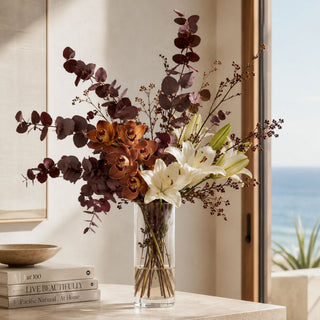 A floral arrangement in a clear glass vase on a tabletop, featuring white lilies, burgundy roses, and dark burgundy eucalyptus stems, with a window and ocean view in the background.