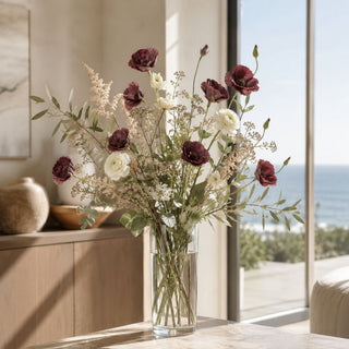 A floral arrangement in a clear glass vase on a table, featuring burgundy bellflowers, white ranunculus, and green foliage, set in a bright room with a window overlooking the ocean.