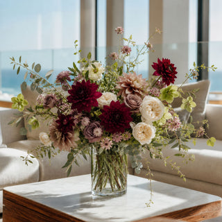 A large, vibrant floral arrangement featuring burgundy, cream, and dusty pink flowers with green foliage, presented in a clear glass vase on a marble-topped table in a bright, modern living room.