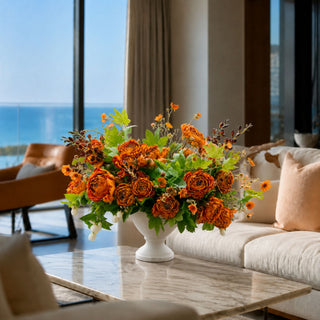 Luxury orange floral arrangement with green foliage in a white pedestal vase, styled on a marble table in an elegant ocean-view California living room for True Bloom Floral.