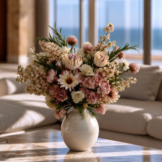 Creamy heirloom mauve king protea faux floral arrangement styled in a rounded ceramic vase with blush roses, ivory florals, soft greenery, and layered botanical texture in a modern living room.