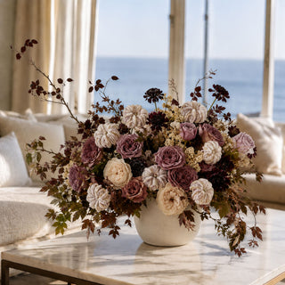 A large floral arrangement in a white ceramic vase sits on a marble coffee table in a bright living room with a window overlooking the ocean.