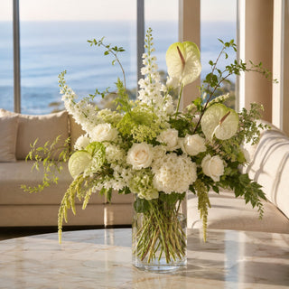 Luxury ivory and green floral arrangement featuring anthuriums, white roses, hydrangeas, and seasonal greenery in a clear glass vase on a marble coffee table overlooking the ocean.