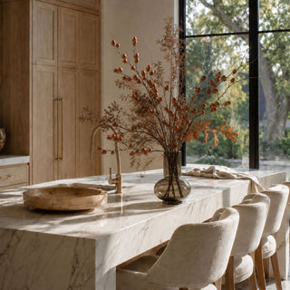 Luxury faux autumn branch arrangement featuring realistic acorn stems, dried botanicals, oak leaves, and airy preserved foliage in a smoked glass vase on a marble kitchen island. Elegant fall home décor styled in a warm modern organic kitchen, ideal for luxury interior design inspiration and seasonal decorating.