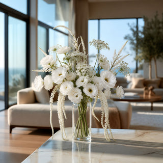 Luxury faux floral arrangement featuring white ranunculus, airy Queen Anne's lace, and cascading white amaranthus stems in a clear glass vase, styled on a marble coffee table in a modern coastal-inspired living room.