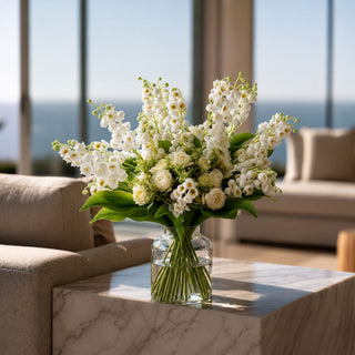 White Real Touch Lily of the Valley floral arrangement styled in a clear glass vase on a modern coffee table with soft white blooms, layered greenery, and elevated year-round interior styling.