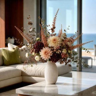 A large dried floral arrangement in a textured cream ceramic vase, set on a marble coffee table in a modern living room with a sofa and a view of the ocean through large windows.