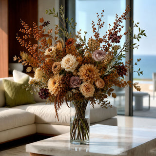 A large dried floral arrangement in a clear glass vase, placed on a marble coffee table in a bright living room with a sofa and large windows overlooking water.