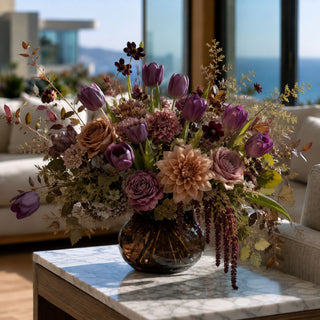 Luxury fall floral arrangement with dark purple real touch tulips, café au lait dahlias, toffee roses, antique hydrangeas and burgundy amaranthus in a smoked amber glass vase on a marble coffee table.