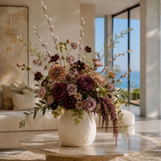 Luxury late summer to early fall faux floral arrangement featuring burgundy velvet willow stems, wine garden roses, taupe dahlias, antique mauve roses, chocolate cosmos, olive branches, and trailing plum amaranthus in a textured white ceramic vase. styled in a bright modern living room.