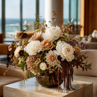 Luxury fall floral arrangement featuring white real touch peonies, café au lait dahlias, toffee roses, antique hydrangeas and burgundy amaranthus in a smoked amber glass vase on a marble coffee table.