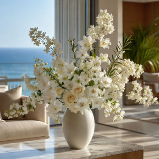 Luxury faux floral arrangement featuring white lily stems, ivory roses, and airy white flowering branches in a matte ivory vase on a marble coffee table in a modern coastal living room overlooking the ocean.