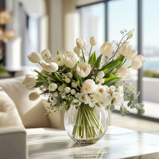 An arrangment of white tulips styled in a clear round vase with greenery and white filler florals on a table in a modern home