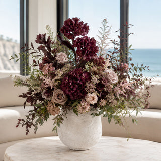 Two-Tone Plum EVA Hydrangea styled in a fall floral arrangement with mauve roses, burgundy blooms, airy filler stems, seasonal greenery, and plum-toned foliage in a textured stone vessel on a marble coffee table in a bright coastal living room.