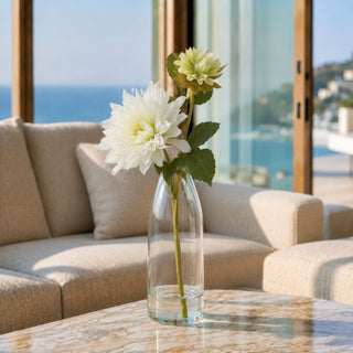 A clear glass vase holding two white dahlias with green stems, placed on a marble table in a bright living room with a beige sofa and large windows overlooking the ocean.