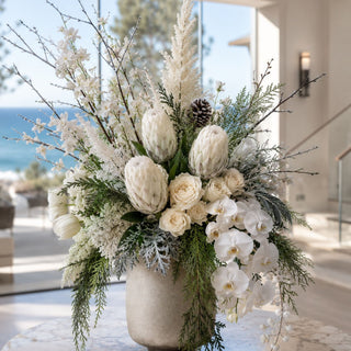 Flocked White 31 Inch King Protea Stem styled in a large winter white floral arrangement on a marble table in a luxury modern home with ocean views
