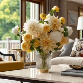 Yellow Real Touch dahlia stems styled in a glass vase with soft cream florals, yellow ranunculus-style blooms, white accents, and green foliage on a coffee table in a warm living room setting.