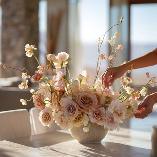 Luxury faux blush rose and soft pastel floral arrangement being styled by hand in a ceramic bowl on a marble table, set in a bright coastal home with warm natural sunlight, elegant high-end home decor