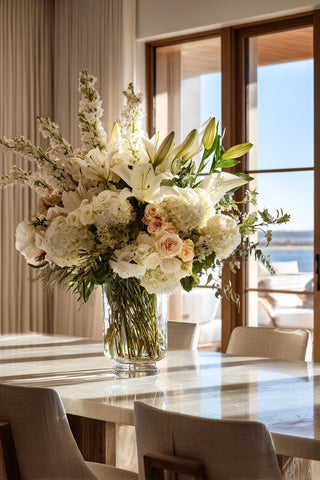 Luxury faux white hydrangea, lily, and rose floral arrangement in a tall glass vase on a marble dining table in a bright modern coastal home with ocean view and natural sunlight, elegant high-end dining room decor