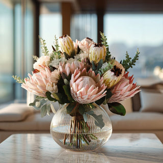 Luxury faux blush protea floral arrangement with greenery in a ribbed glass vase on a marble table, styled in a bright modern coastal home with soft natural light, elegant high-end home decor