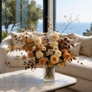 A dried floral arrangement in a clear ribbed glass vase, placed on a light marble coffee table in front of a cream sofa with a window overlooking trees and the ocean.